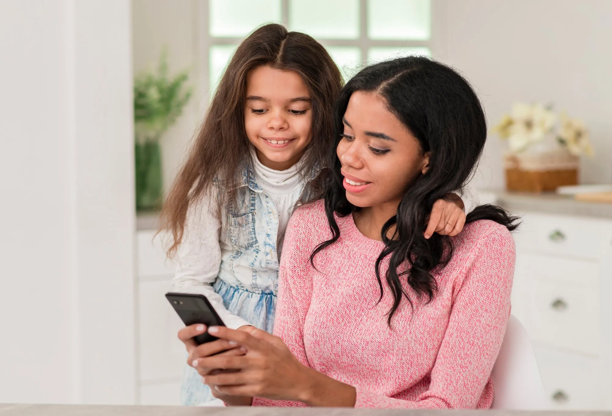 Una madre y su hija miran juntas la pantalla del celular en la cocina de su casa
