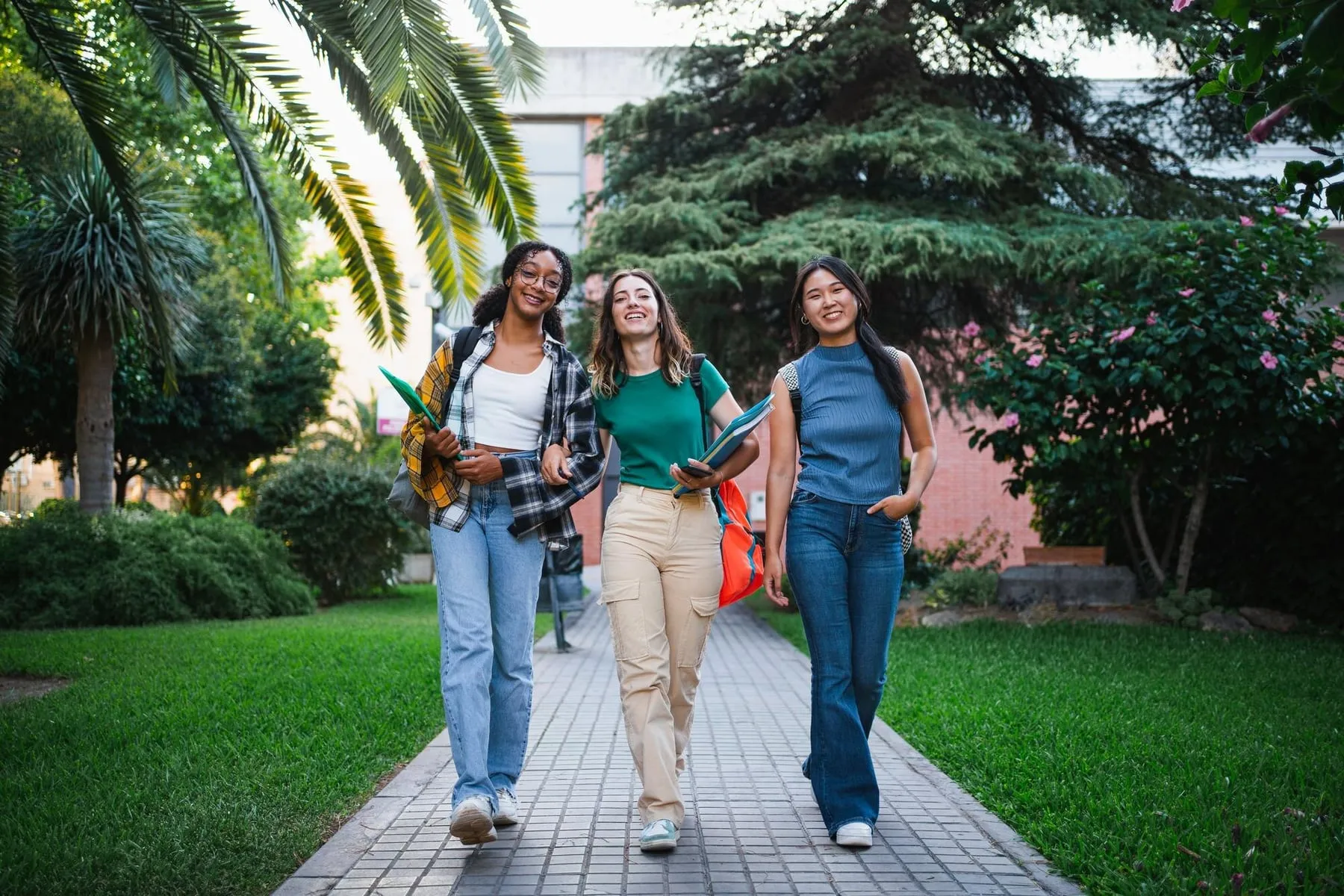 Tres estudiantes universitarias caminan sonrientes por un sendero del campus con sus cuadernos