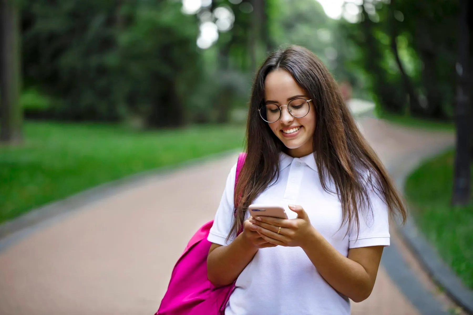 Una estudiante con lentes sonríe mientras escribe en su celular en un sendero del campus